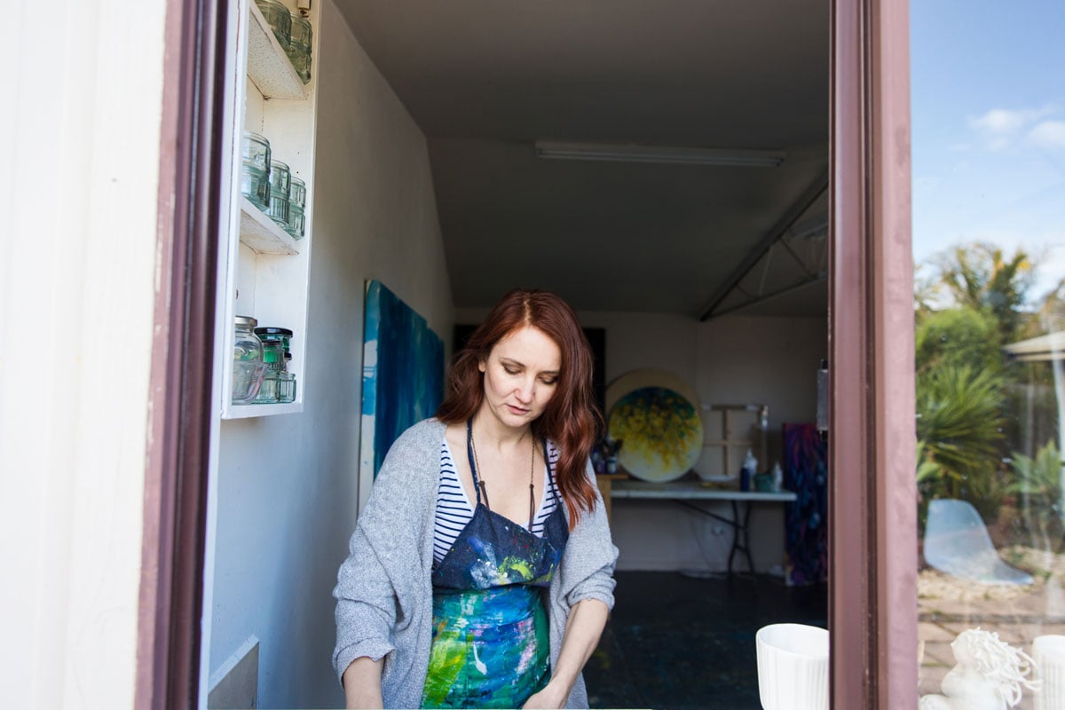 red haired artist washing brushes in the sink, shot through the window of her studio