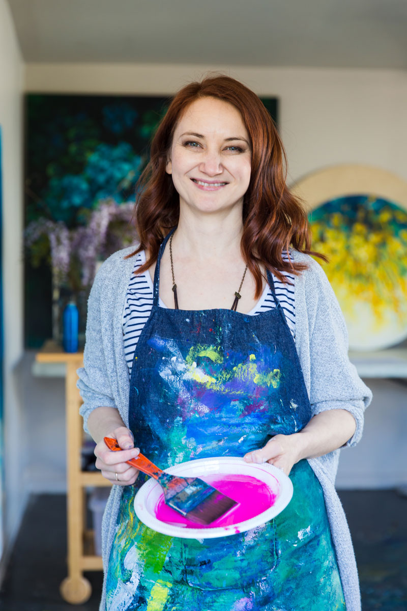 artist holding plastic plate with magenta paint on it, with paintings out of focus in the background