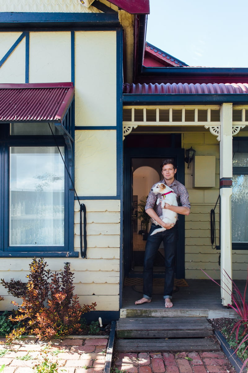 mid-30s man standing on porch of yellow weatherboard home with burgundy and navy details 