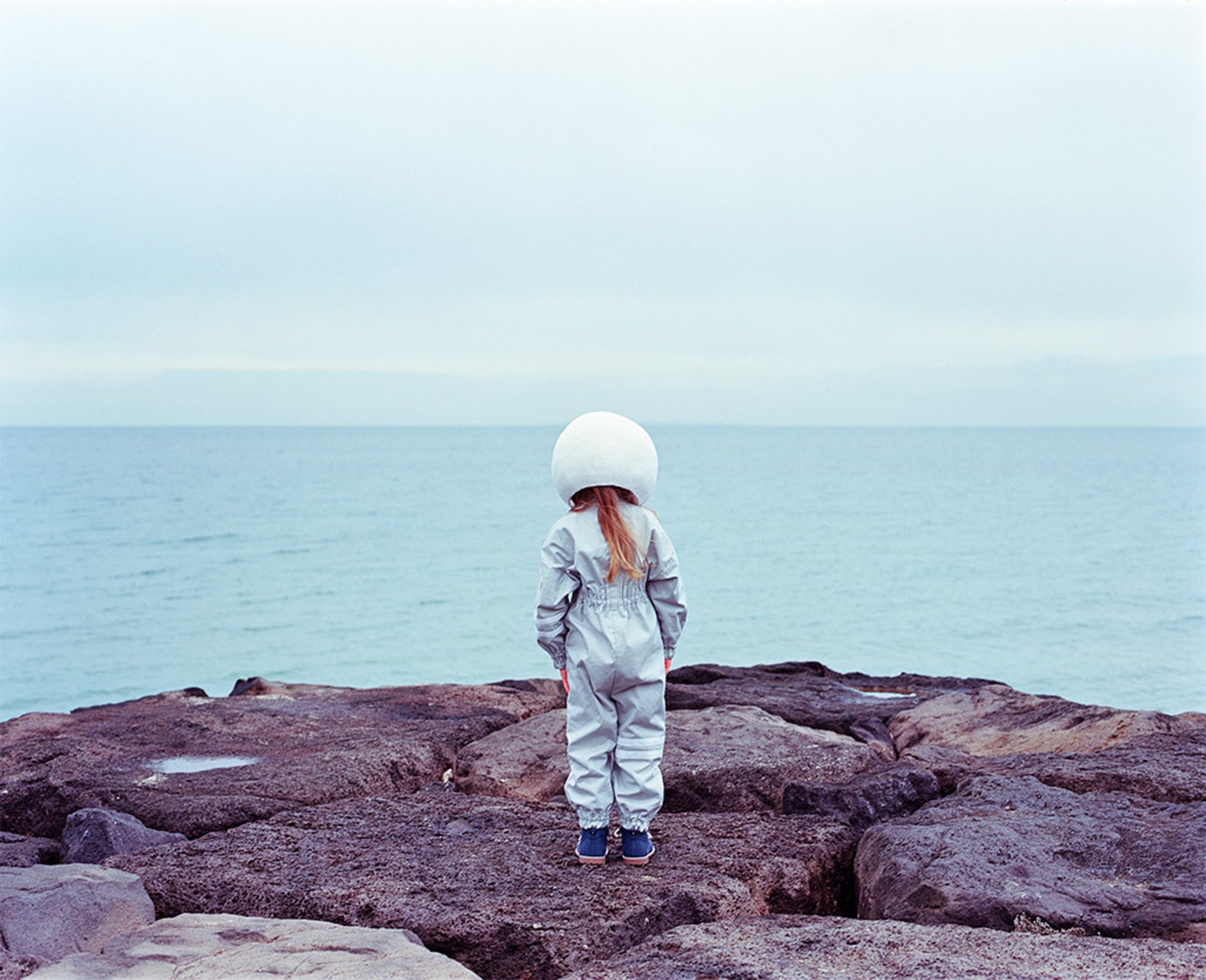 Rocky Shores by Andrew Rovenko - a girl is turned towards the see with her back to the camera in an astronaut suit.
